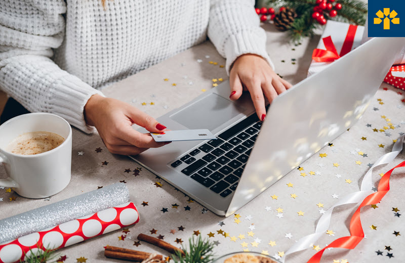 A person is holding a credit card at a laptop. The scene has holiday related decorations and wrapping paper. 
