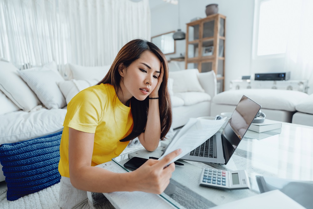 A woman sits on the floor at the coffee table in her living room. She leans over a laptop and calculator with papers in her hand.